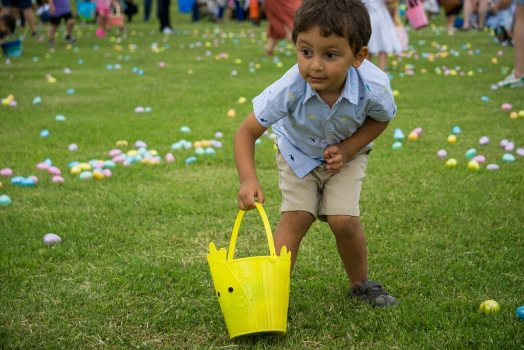 A young boy in a blue shirt and khaki shorts bends down holding a yellow bucket on a grassy field scattered with colorful plastic eggs, participating in an outdoor egg hunt. Other people and children are in the background.