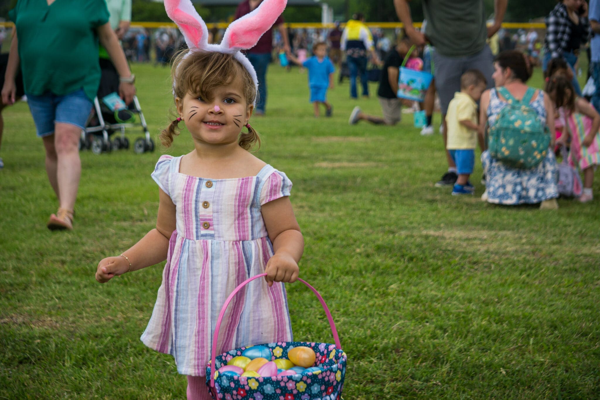 A young girl wearing bunny ears and whiskers, holding a colorful basket filled with Easter eggs, smiles while standing on grass at an outdoor event with people in the background.