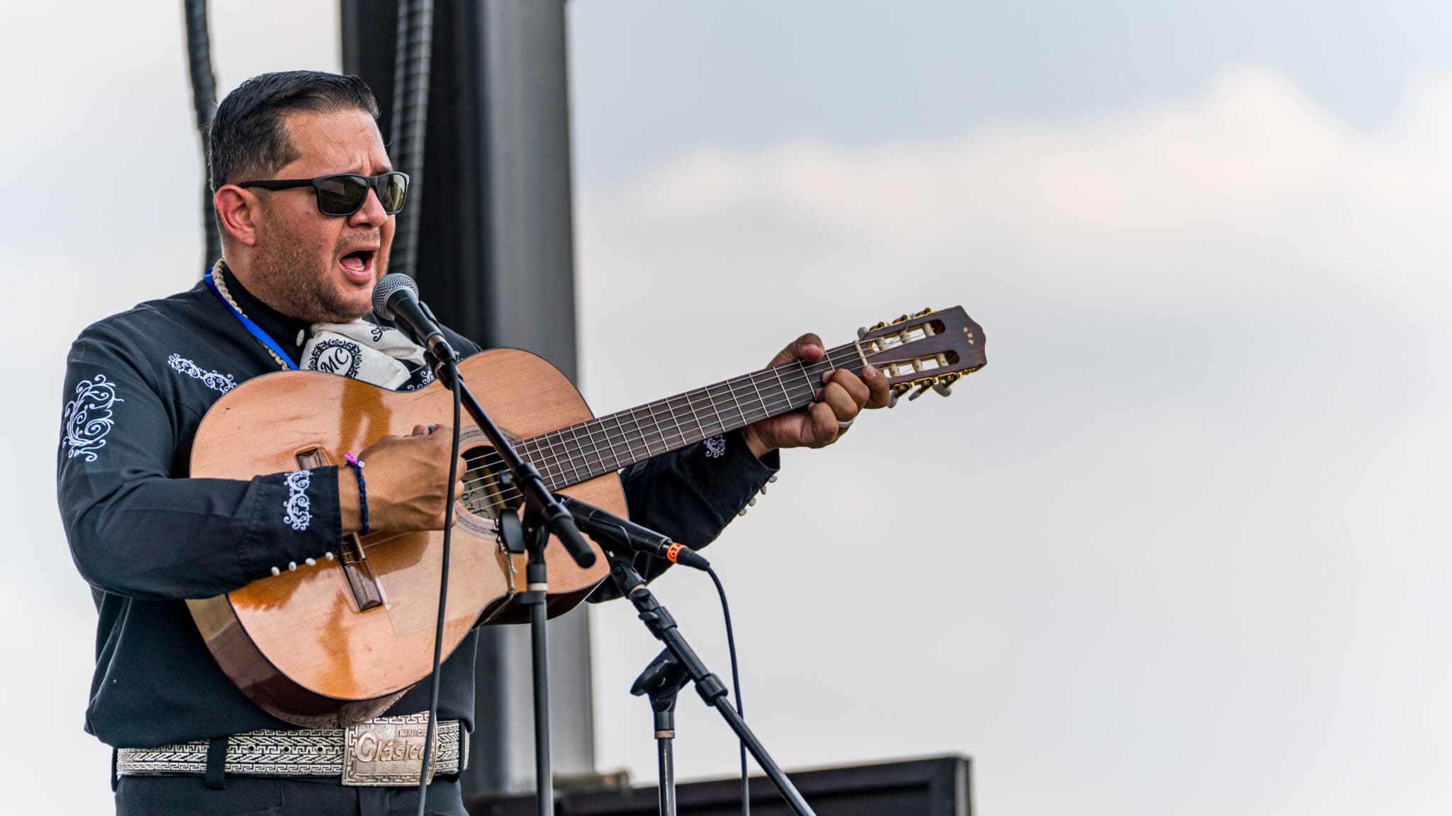 A man wearing sunglasses and a black embroidered shirt passionately sings and plays an acoustic guitar on stage, with microphones set up in front of him and a cloudy sky in the background.