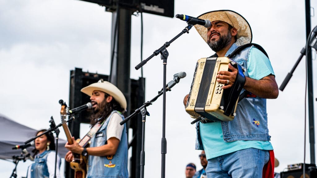 Three musicians perform on an outdoor stage. One sings and plays the accordion, another sings and plays guitar, and a third sings in the background. All wear cowboy hats and denim vests. The sky is cloudy.