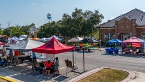 A street market scene with colorful tents and booths set up along both sides of a road, people browsing and working at the stalls, and a brick building with a clock tower in the background on a sunny day.