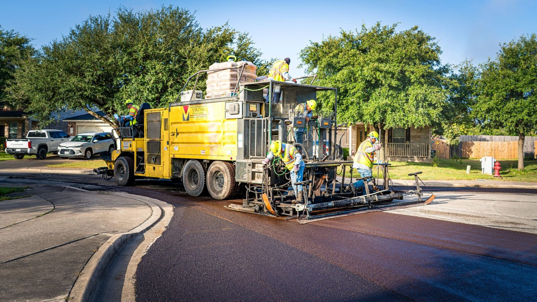 A yellow road paving machine with several workers in safety gear resurfacing a residential street, surrounded by trees and houses on a sunny day.