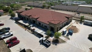 Aerial view of a large, single-story building with a red tile roof, surrounded by a parking lot with several cars and construction materials nearby. Trees and other buildings are visible in the background.