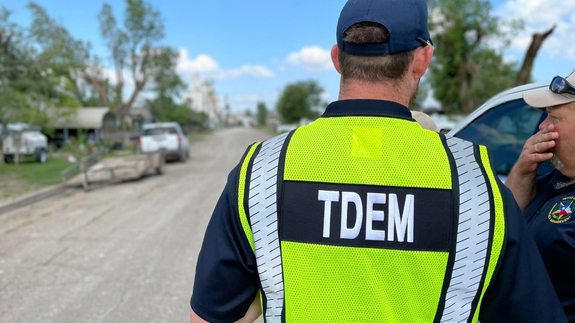 A person wearing a yellow safety vest labeled "TDEM" stands on a dirt road, facing away, next to another person in uniform. Several vehicles and damaged trees are visible in the background.