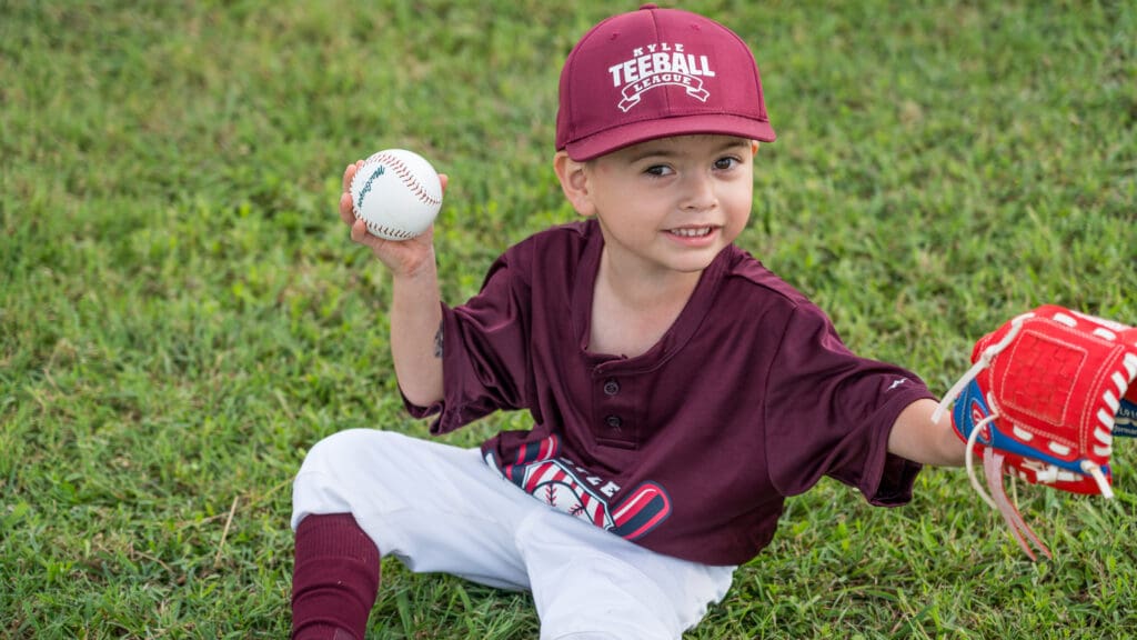 A young child in a maroon baseball uniform and cap sits on grass, smiling, holding a baseball in one hand and a red glove in the other. as part of the Kyle Tee-Ball League.