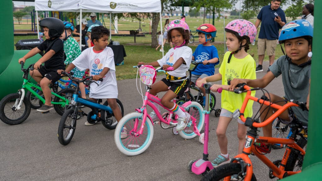 A group of young children wearing helmets and casual clothes are gathered with their bikes and a scooter at a starting line outdoors, preparing for a race or event. Adults and tents are visible in the background.