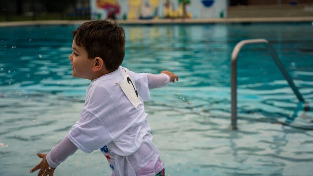 A young boy in a white shirt with a paper number pinned on the back stands in a swimming pool, arms outstretched, smiling as water splashes around him. A metal pool ladder and colorful mural are visible in the background.