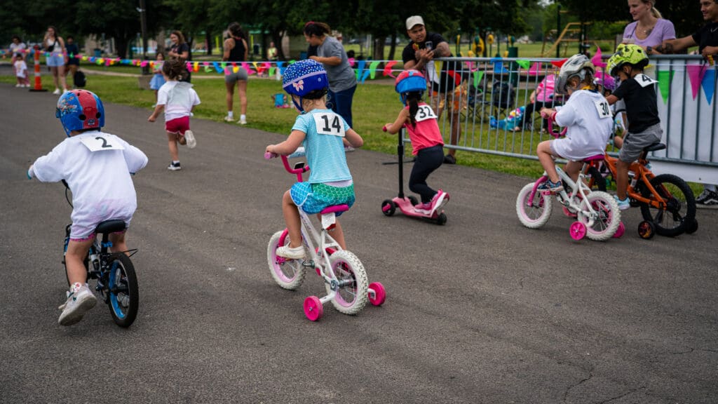 Young children wearing helmets and numbered shirts ride bikes and scooters in a race at a park, while adults and other kids watch from behind a fence decorated with colorful flags.