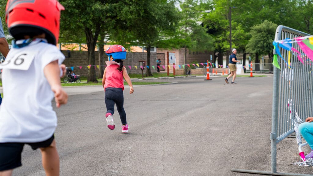 Young children wearing helmets and numbered shirts run down a paved street during an outdoor race, with colorful flags and trees in the background. An adult and another child watch from the sidelines.