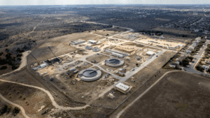 Aerial view of a large construction site with several circular and rectangular structures, surrounded by dirt roads, open land, and nearby residential neighborhoods.