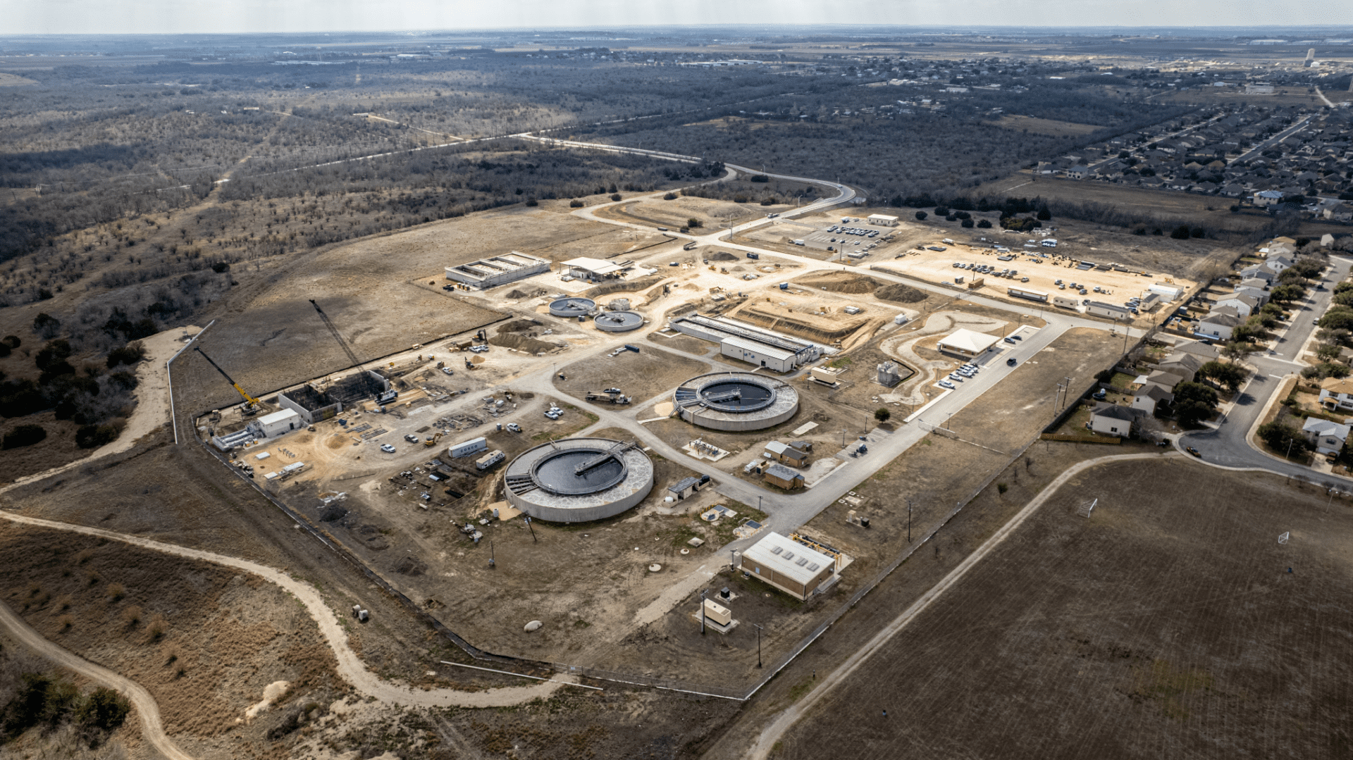 Aerial view of a large construction site with several circular and rectangular structures, surrounded by dirt roads, open land, and nearby residential neighborhoods.