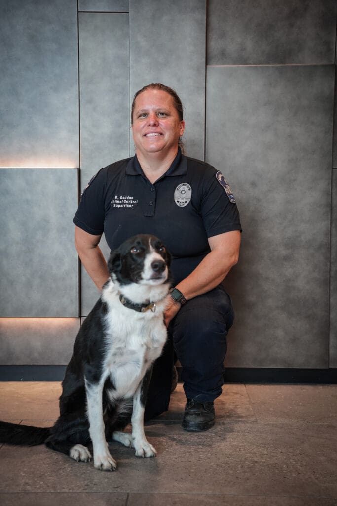 A person in a dark uniform kneels next to a black and white dog indoors, both facing the camera and posing in front of a modern, gray tiled wall.