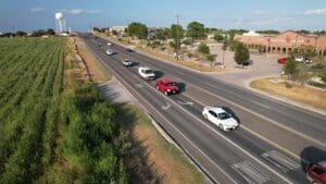 An aerial view of a multi-lane road with several cars driving in both directions, bordered by a field on the left and businesses with parking lots on the right; a water tower is visible in the background.