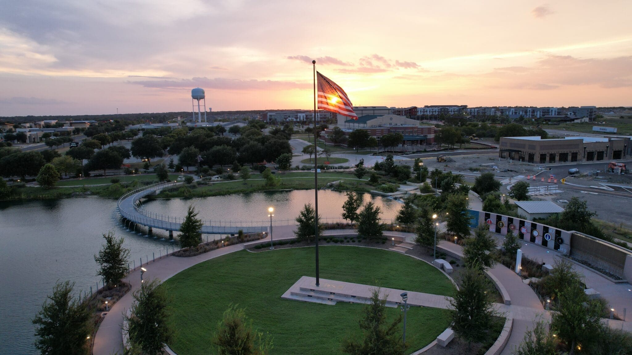A large American flag flies in the center of a circular green park by a pond at sunset, with buildings, a bridge, trees, and a water tower visible in the background.
