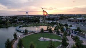 A large American flag flies in the center of a circular green park by a pond at sunset, with buildings, a bridge, trees, and a water tower visible in the background.