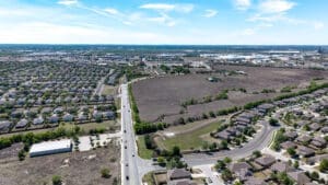 Aerial view of a suburban area with houses, roads, and a large empty field in the center. The landscape extends to the horizon under a bright blue sky with scattered clouds.