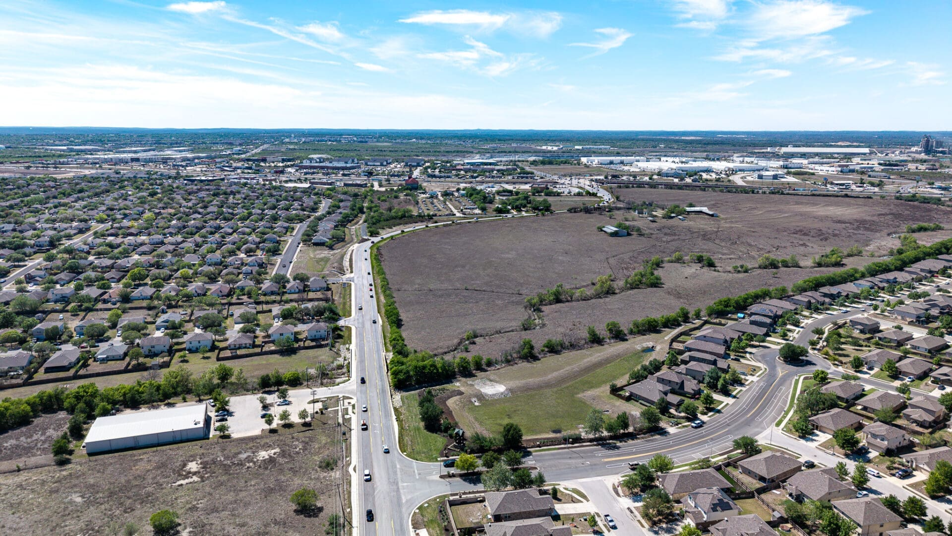 Aerial view of a suburban area with houses, roads, and a large empty field in the center. The landscape extends to the horizon under a bright blue sky with scattered clouds.