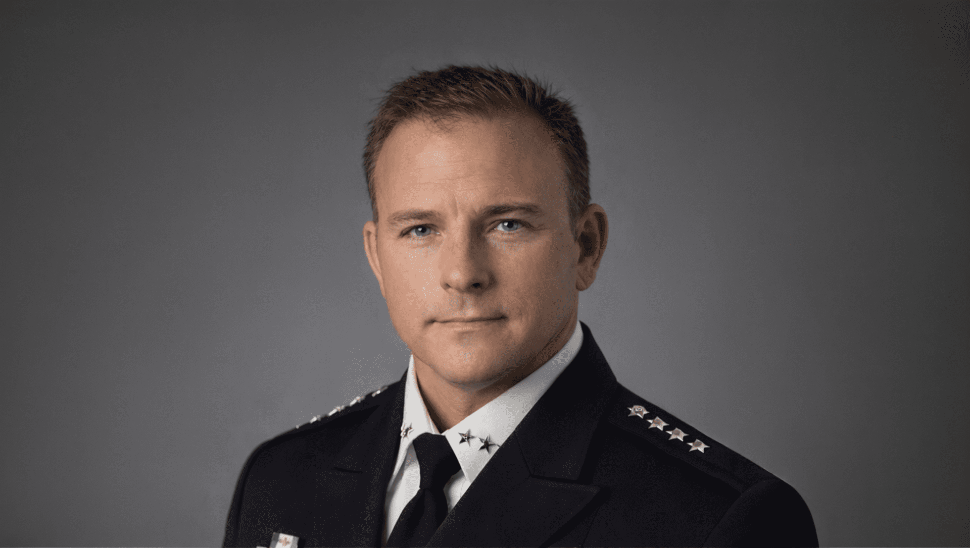 A man in a dark police or military uniform with four stars on each shoulder poses against a plain gray background, looking directly at the camera with a neutral expression.