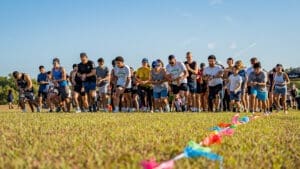 A large group of people in athletic wear line up at the starting line of a race on a grassy field, preparing to run. Colorful flags mark the starting line under a clear blue sky.