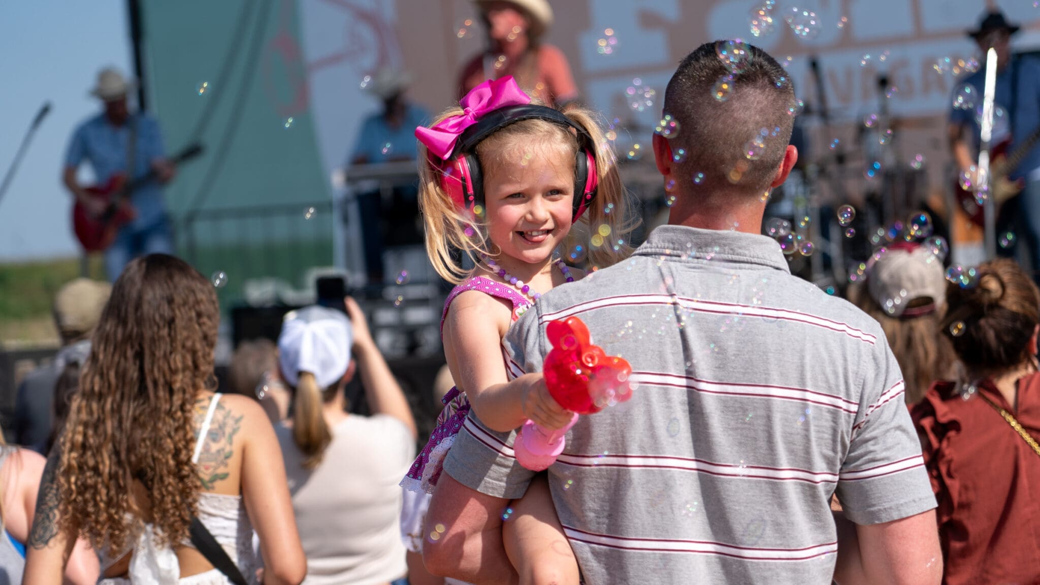 A man holds a smiling young girl with pink headphones and a balloon animal at an outdoor concert, surrounded by bubbles and a crowd on a sunny day.