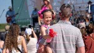A man holds a smiling young girl with pink headphones and a balloon animal at an outdoor concert, surrounded by bubbles and a crowd on a sunny day.