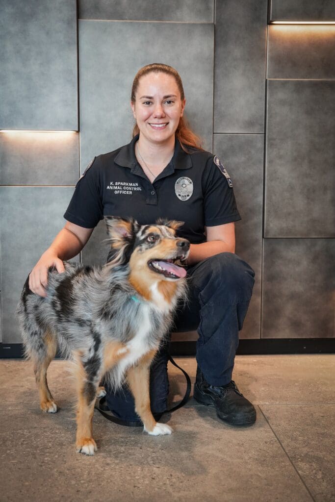 A smiling woman in a dark uniform crouches indoors next to a happy Australian Shepherd dog with a merle coat. The background features modern gray wall panels.
