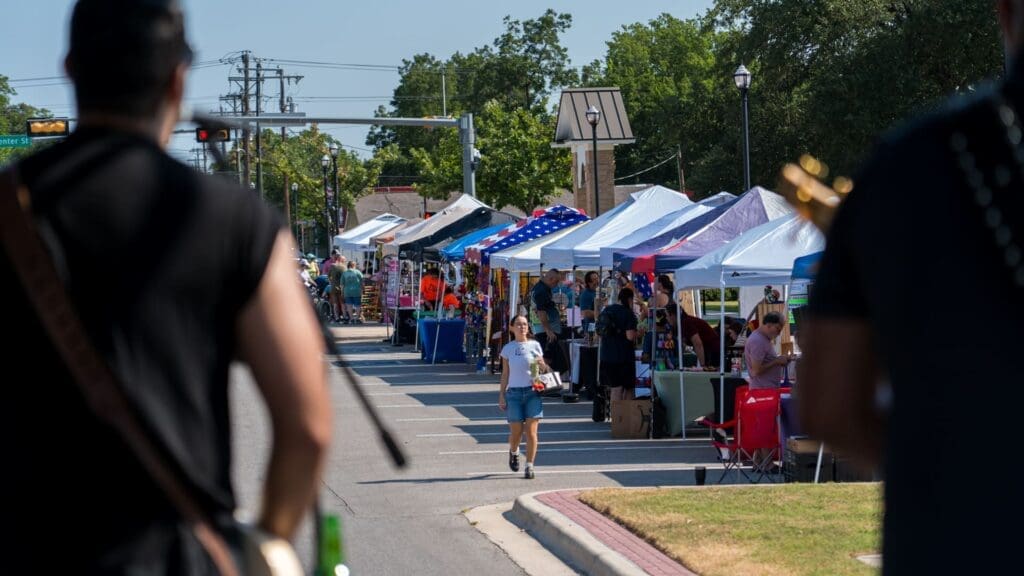 A street fair with colorful vendor tents lined up on both sides of the street. A person walks down the road carrying items, while two musicians play in the foreground. Trees and a few buildings are visible in the background.