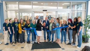 A group of people stand in front of glass doors cutting a large blue ribbon at a ceremony. Several hold the ribbon while others clap and smile, and a few people wear matching shirts with a logo.