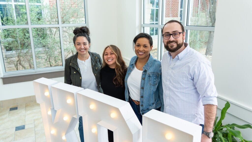 Four smiling people stand together indoors by large lit-up letters, next to tall windows with trees visible outside.