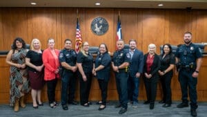 A group of eleven people, including police officers in uniform and civilians in business attire, stand side by side in front of wooden paneling, an American flag, and a city seal inside a courtroom or council chamber.