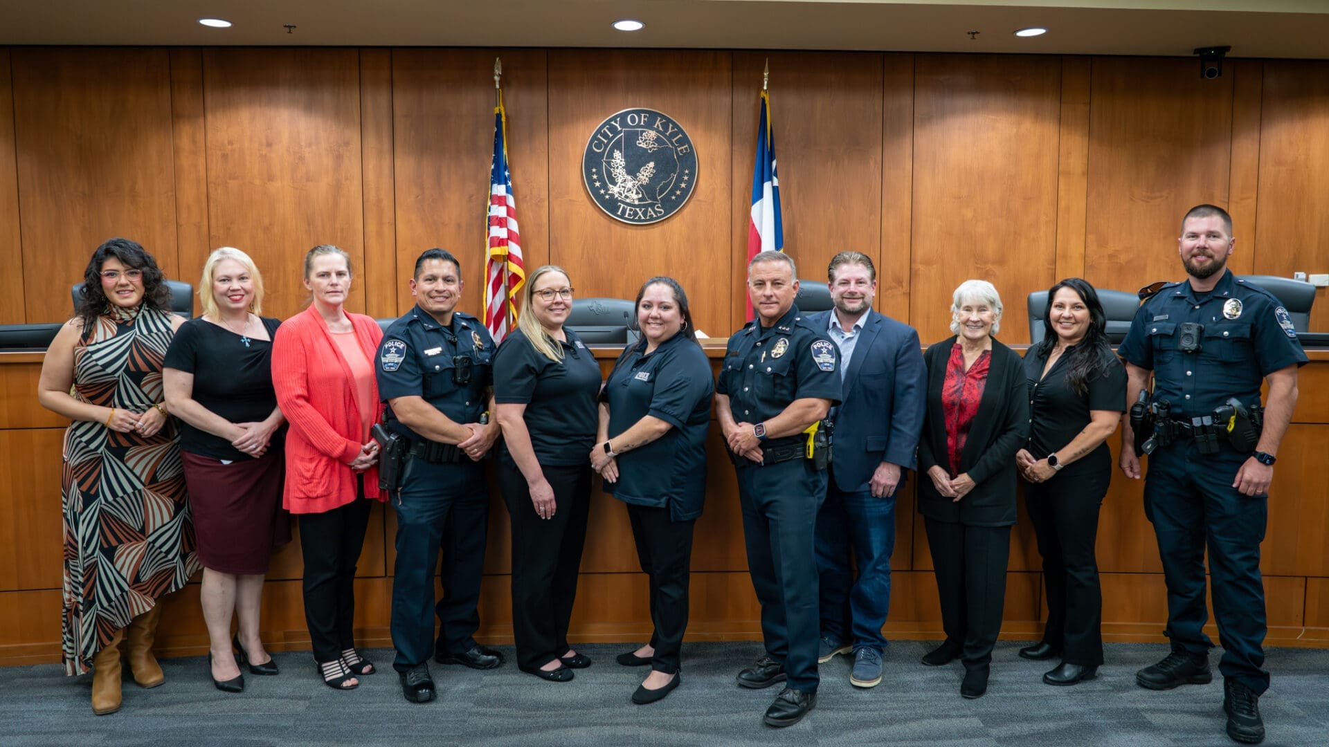 A group of eleven people, including police officers in uniform and civilians in business attire, stand side by side in front of wooden paneling, an American flag, and a city seal inside a courtroom or council chamber.