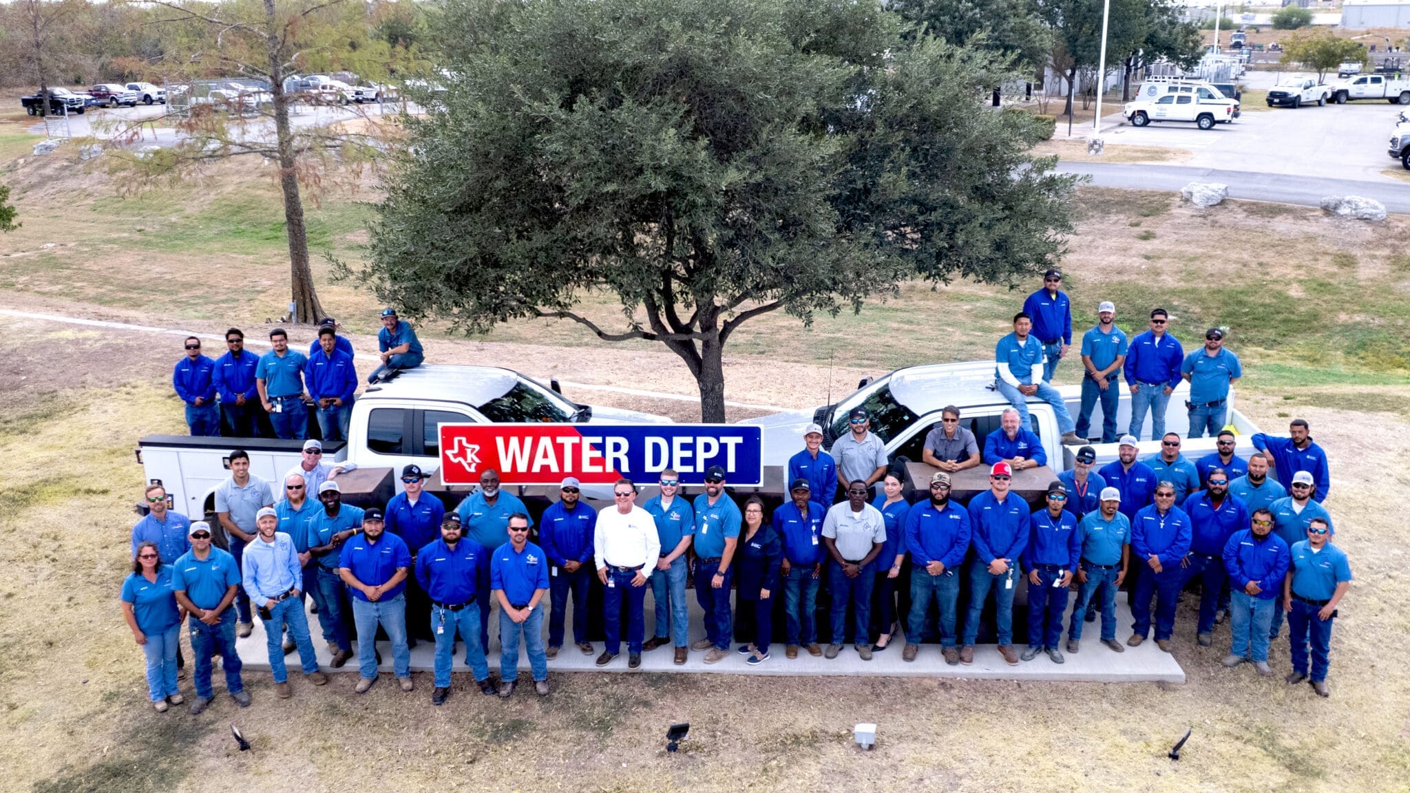 A large group of water department employees wearing blue uniforms stands outdoors in front of trucks and a tree, posing with a sign that reads "WATER DEPT.