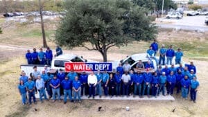A large group of water department employees wearing blue uniforms stands outdoors in front of trucks and a tree, posing with a sign that reads "WATER DEPT.