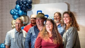 Six adults stand together smiling in front of a city sign and a festive balloon arrangement featuring blue, gold, and red balloons; the group appears to be at an indoor event.