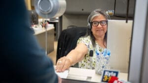 A smiling woman wearing glasses and a headset sits at a desk behind a glass partition, reaching out to take a document from a person standing on the other side. Office supplies and a nameplate are visible on her desk.