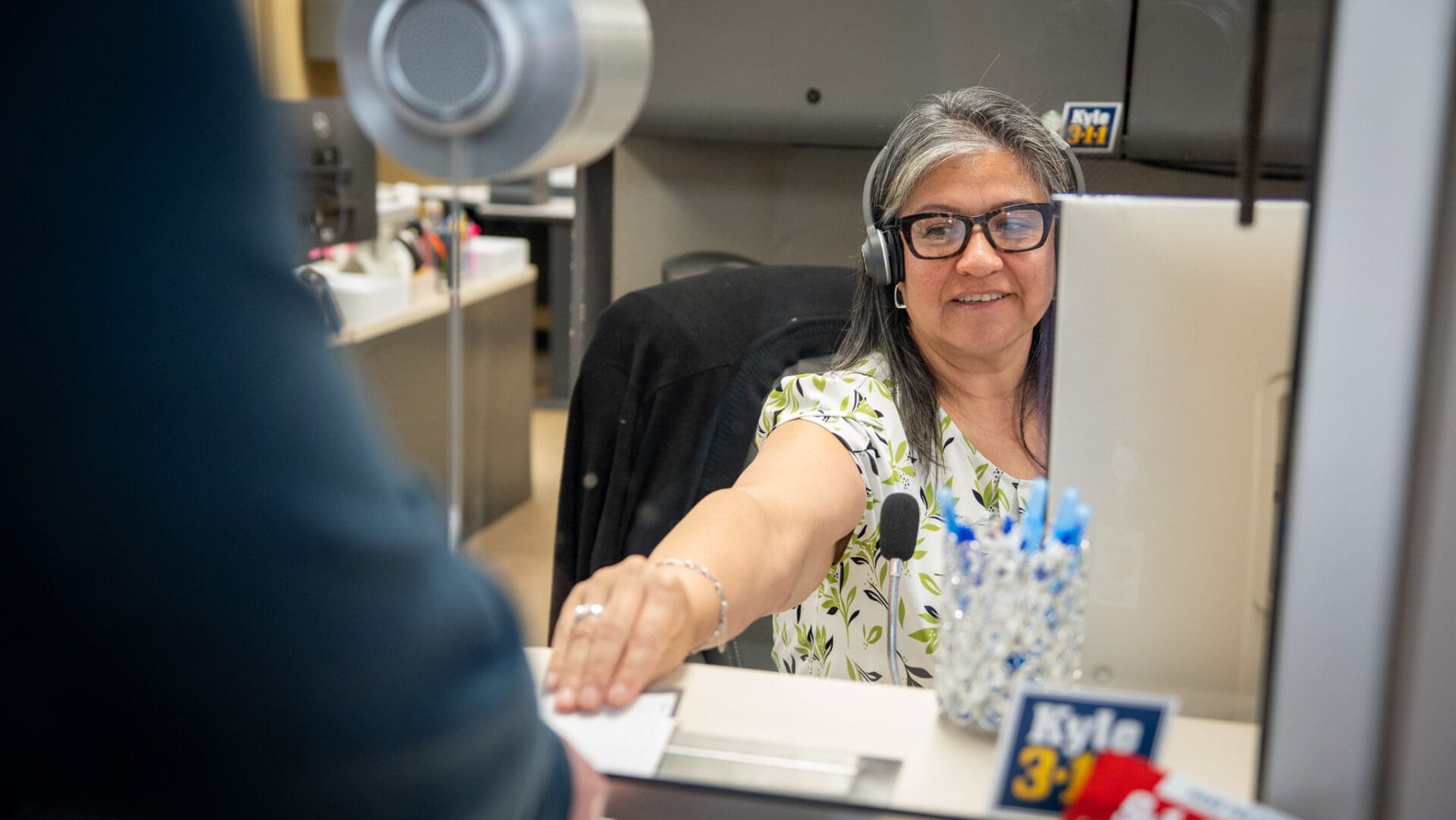 A smiling woman wearing glasses and a headset sits at a desk behind a glass partition, reaching out to take a document from a person standing on the other side. Office supplies and a nameplate are visible on her desk.