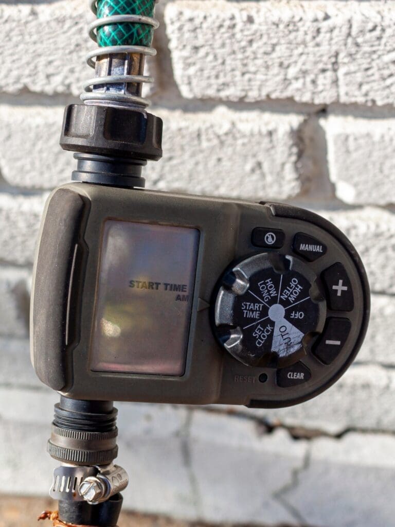 A close-up of an outdoor digital water timer attached to a hose, mounted on a brick wall. The timer's screen displays "START TIME," and the control dial and buttons are visible.