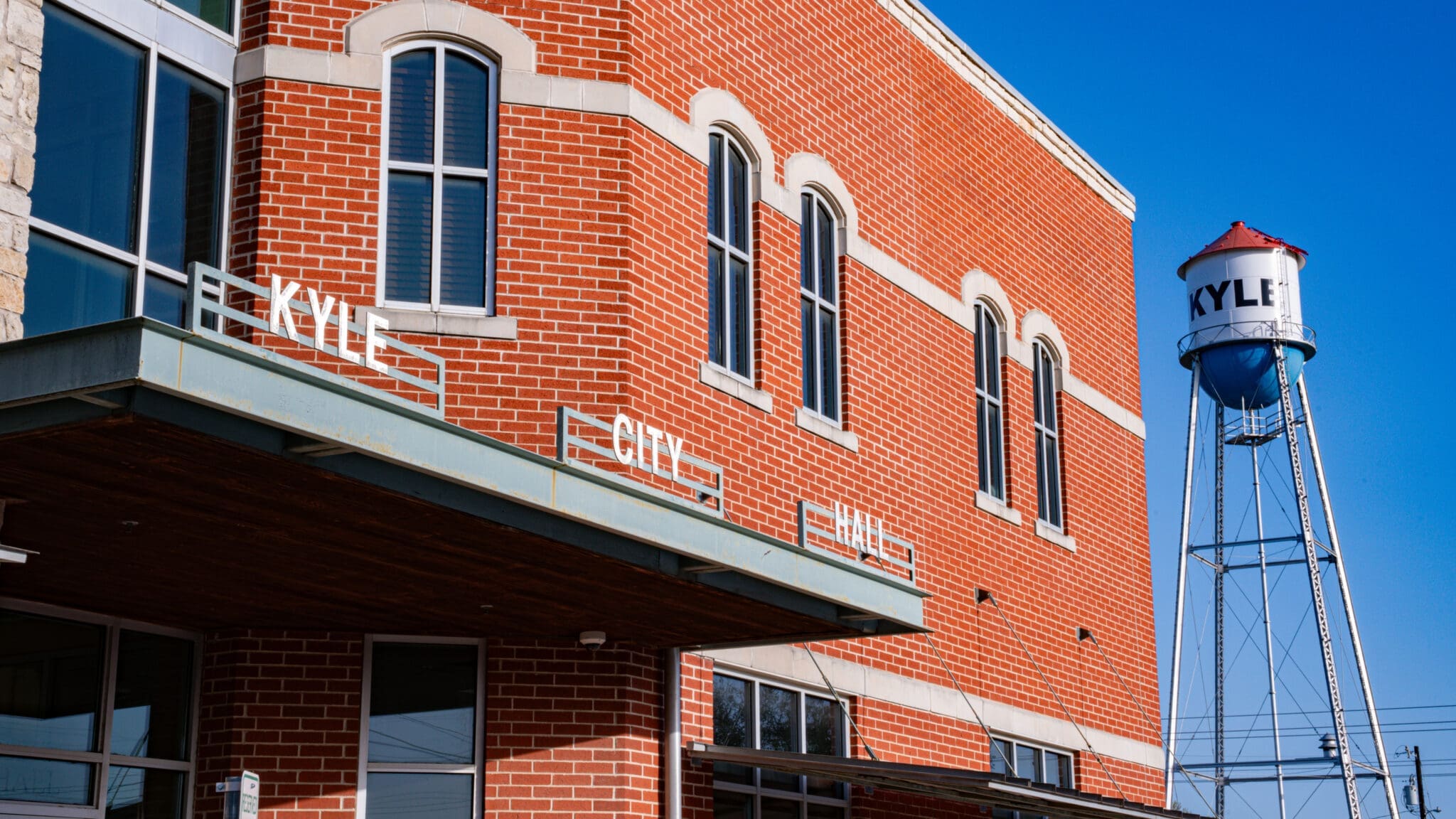 A red brick building labeled "Kyle City Hall" stands in the foreground, with a tall water tower labeled "Kyle" visible against a clear blue sky in the background.