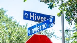 Two blue street signs at an intersection read "Hiver St" and "Invierno St" against a background of green trees and a clear sky. Part of a red stop sign is visible below the street signs.