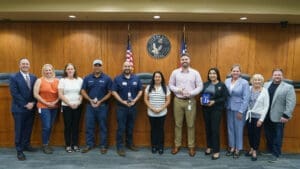 A group of eleven people stand side by side in a government chamber, some holding awards. Behind them are wood-paneled walls, two flags, and the City of Katy, Texas seal. Everyone is smiling for the photo.