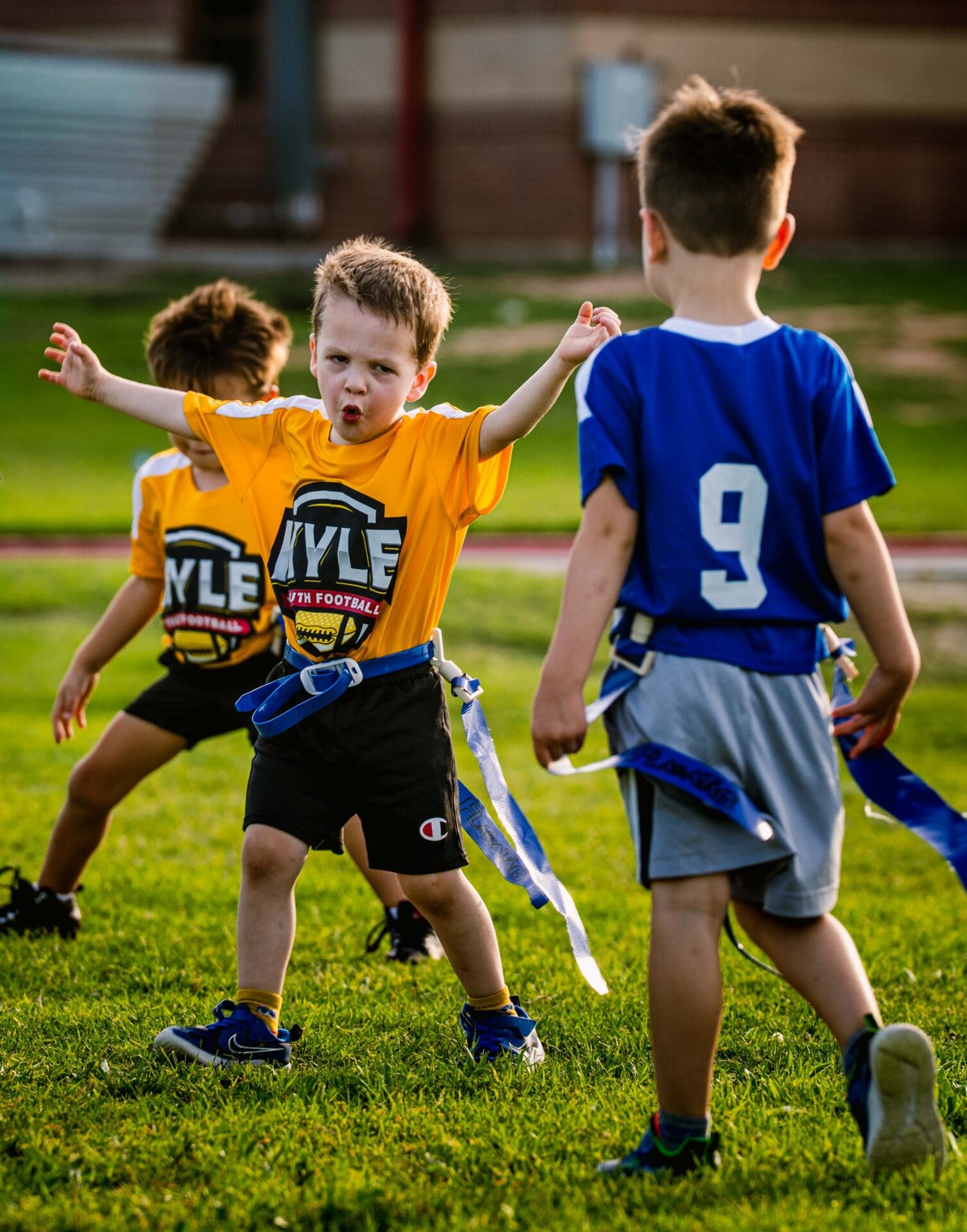Young boy in a yellow jersey excitedly spreads his arms during a youth flag football game, facing another child in a blue jersey with the number 9. Two other kids are visible on the green grass field.