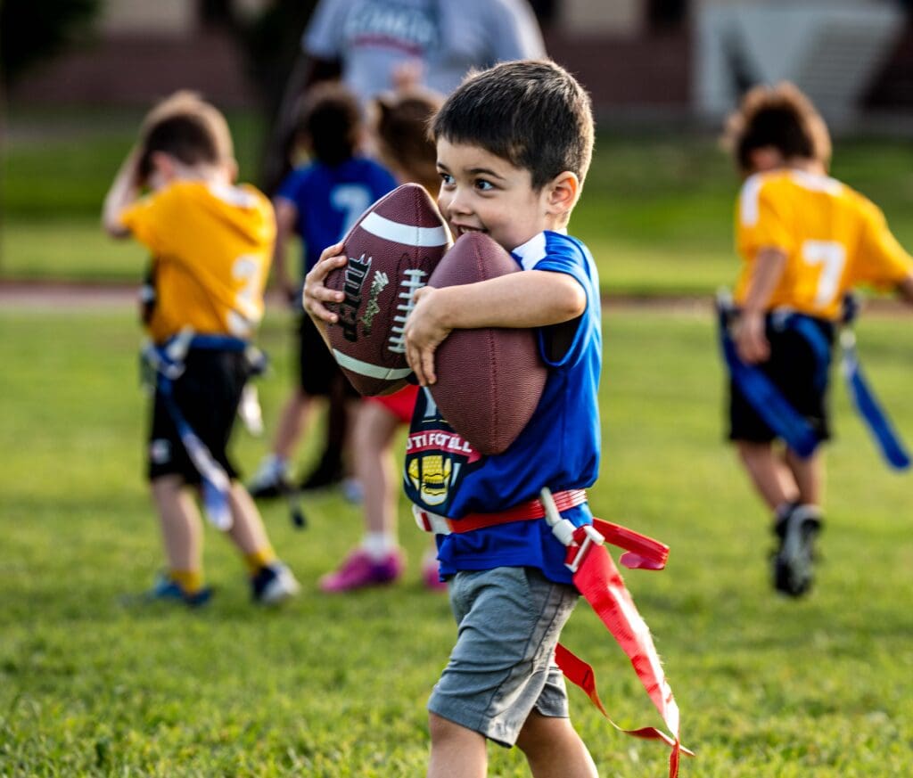 A young boy in a blue shirt and shorts smiles as he hugs a football during a flag football game, with other children in yellow jerseys playing in the background on a grassy field.