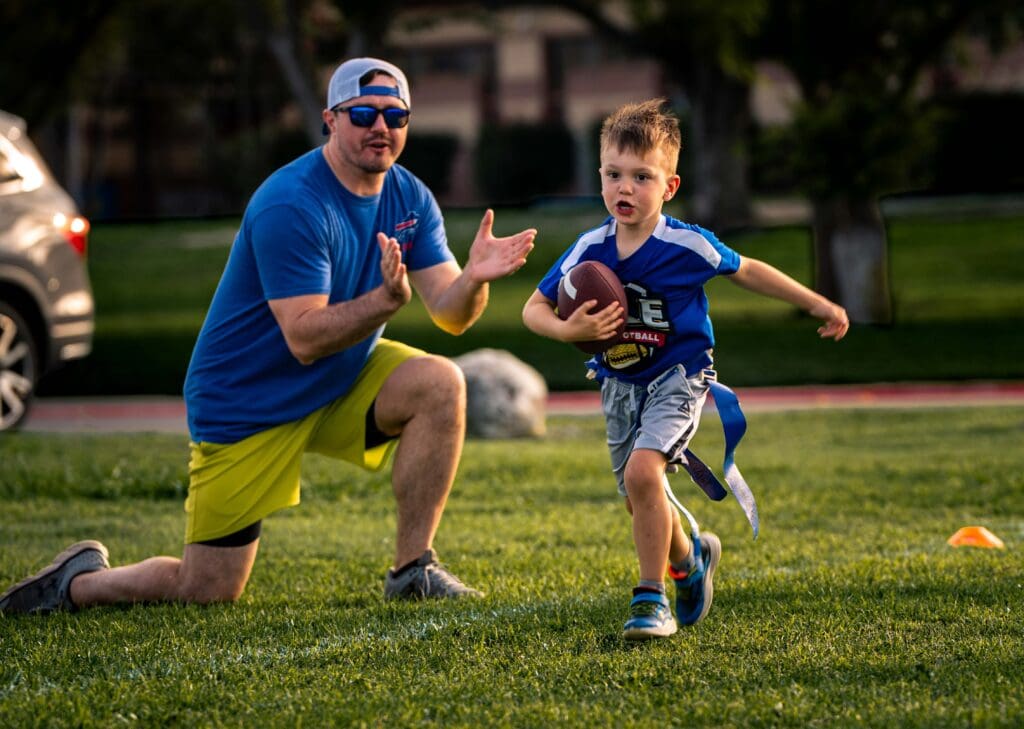 A young boy runs with a football while a man kneels nearby, clapping and cheering him on. They are outdoors on a grassy field with cones, suggesting a youth football or flag football practice.