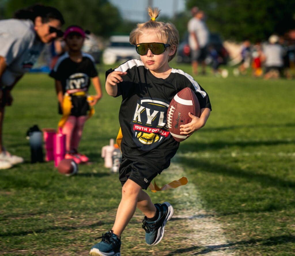 A young child wearing sunglasses and a "Kyle Youth Football" shirt runs on a grassy field holding a football, with other children and adults in the background during a game.