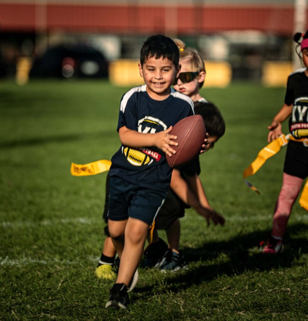 A young boy smiles while running with a football during a flag football game, with other children chasing him on a grassy field.