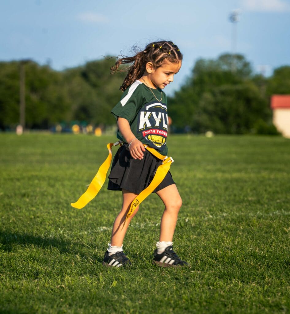 A young child with braided hair wearing a "Kyle Youth Football" jersey and black shorts walks on a grassy field, holding yellow flag football belts. Trees and a blue sky are in the background.