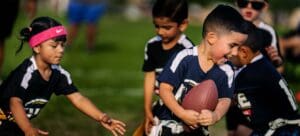 A group of young children play flag football outdoors. One boy clutches a football and runs while a girl wearing a pink headband reaches for his flag. Other children watch and participate in the game on a grassy field.