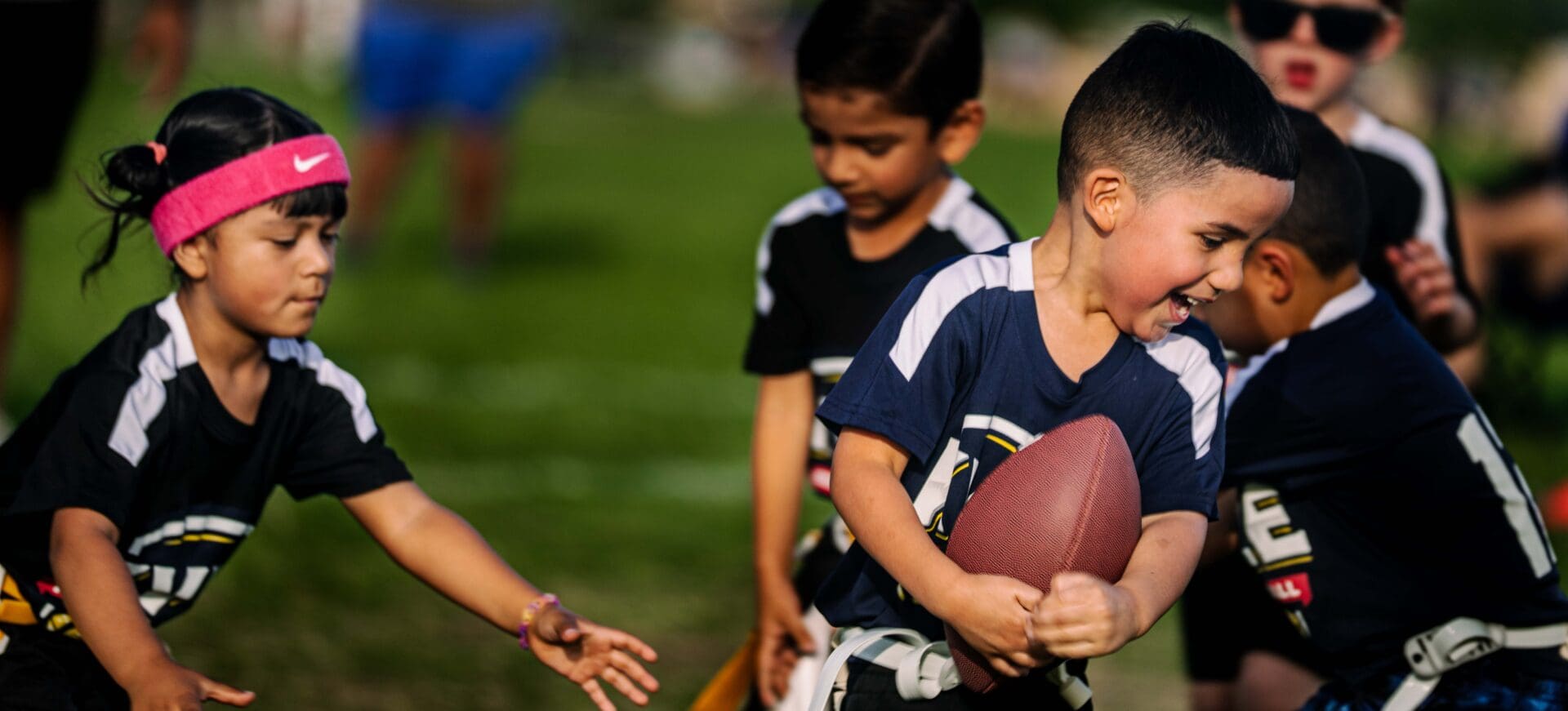 A group of young children play flag football outdoors. One boy clutches a football and runs while a girl wearing a pink headband reaches for his flag. Other children watch and participate in the game on a grassy field.