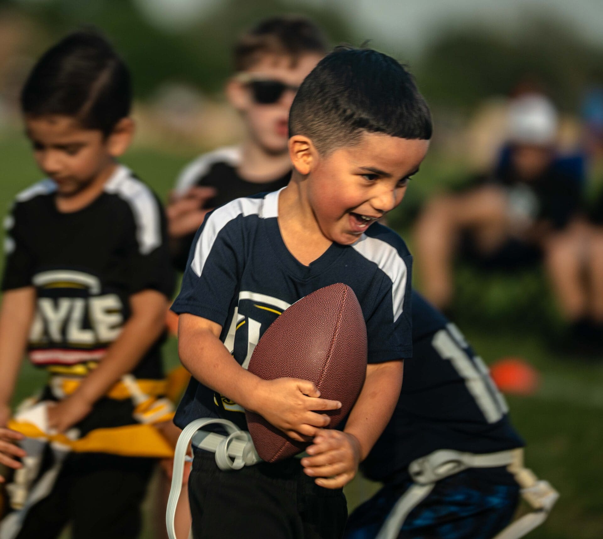 A young boy smiles while running with a football during a youth flag football game, with other children and blurred spectators in the background.