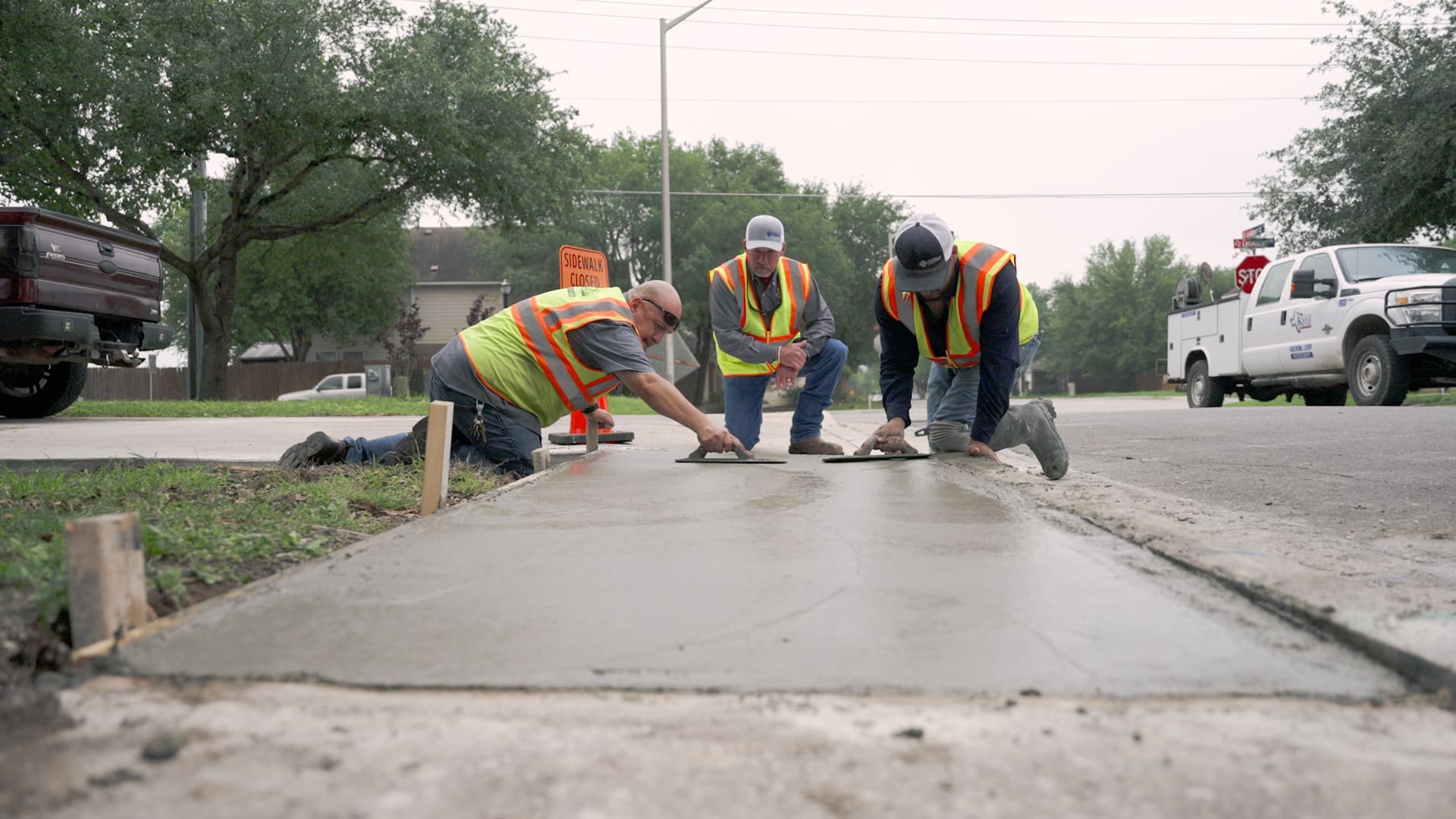 Three construction workers in safety vests and hats smooth freshly poured concrete on a sidewalk near a street, with vehicles and trees visible in the background.